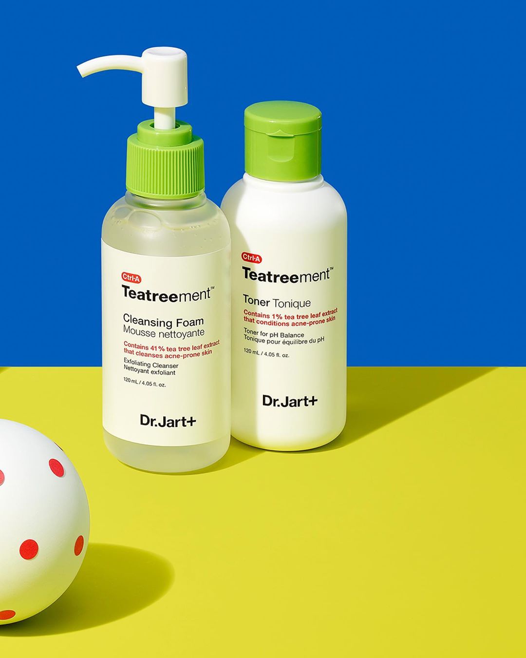Two Dr. Jart+ Teatreement skincare bottles, a cleansing foam and a toner, are displayed on a yellow surface against a blue backdrop.