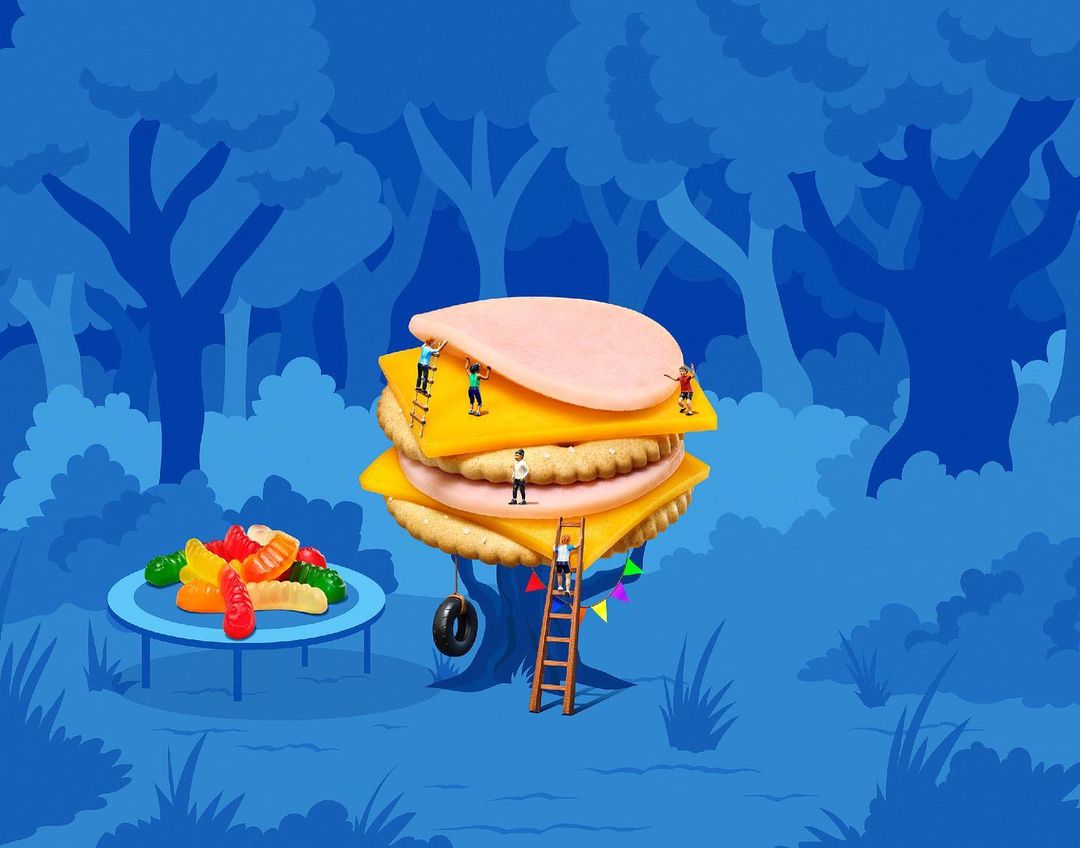 Whimsical food art of a cracker sandwich with cheese and bologna surrounded by tiny people and gummy worms in a blue forest.