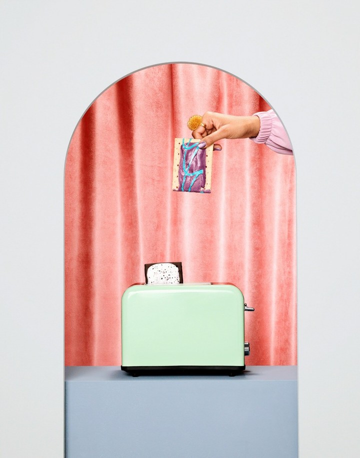 A light green toaster sits on a grey surface in front of a pink curtain, as a hand holds a snack above the toaster.