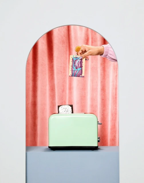 A light green toaster sits on a grey surface in front of a pink curtain, as a hand holds a snack above the toaster.