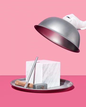 A white-gloved hand lifts a silver cloche dome revealing a marble cube, metal rod, and small hammer on a silver tray against pink background.