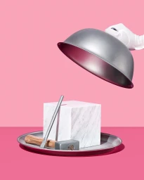 A white-gloved hand lifts a silver cloche dome revealing a marble cube, metal rod, and small hammer on a silver tray against pink background.