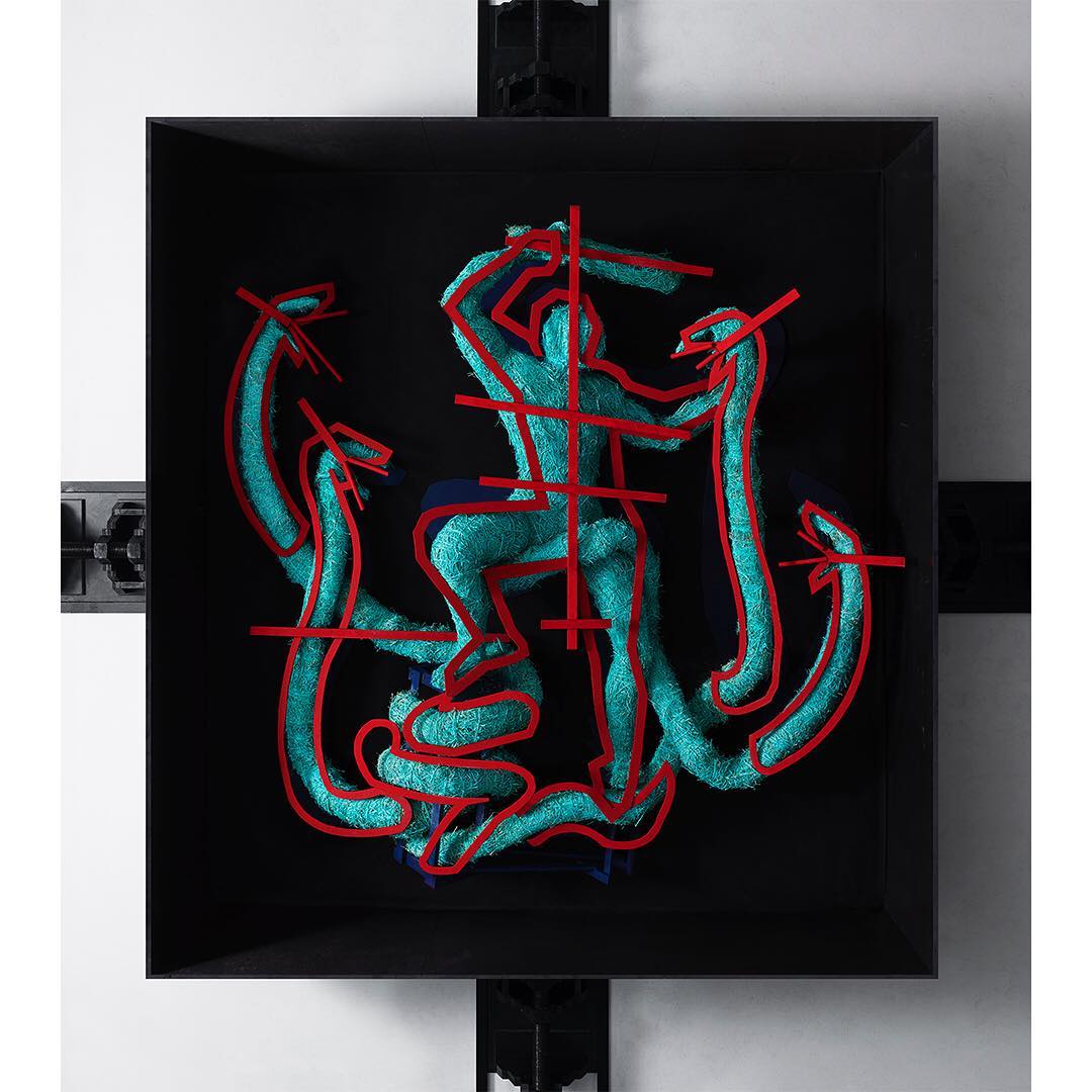 Abstract art in a black frame featuring a turquoise figure outlined in red, riding snakes in an indoor setting against a white wall.