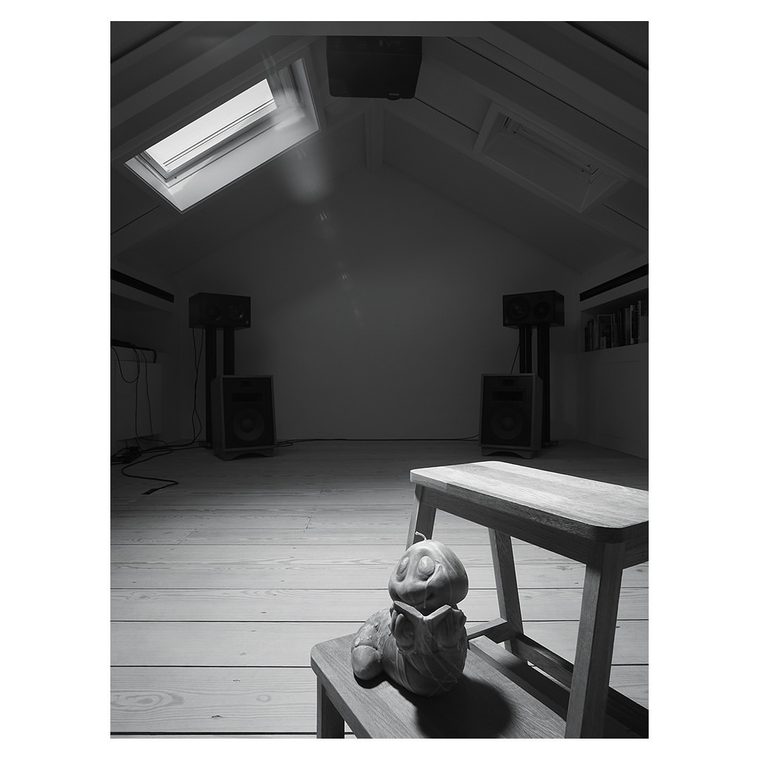 Monochrome shot of a pumpkin figurine sitting on a wooden stool in a room with speakers and natural light from a skylight.