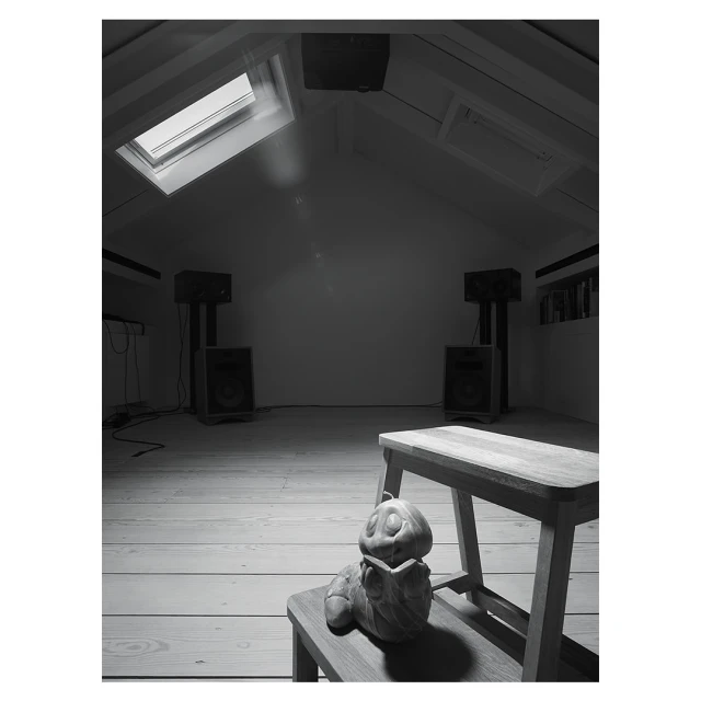 Monochrome shot of a pumpkin figurine sitting on a wooden stool in a room with speakers and natural light from a skylight.