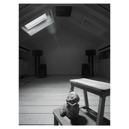 Monochrome shot of a pumpkin figurine sitting on a wooden stool in a room with speakers and natural light from a skylight.