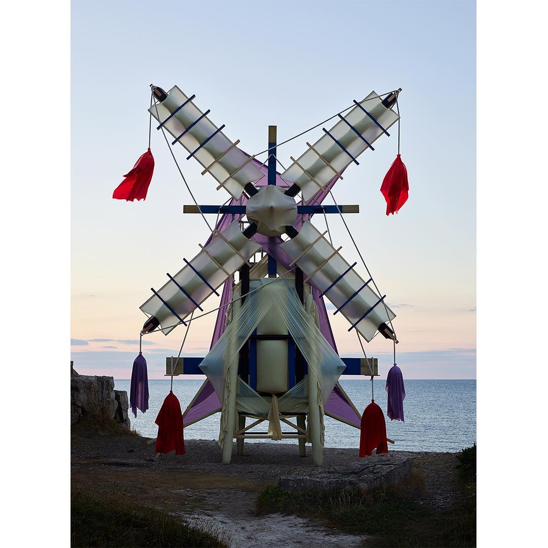 Art installation of a windmill structure with colorful fabric accents, set against a backdrop of the sea and evening sky.