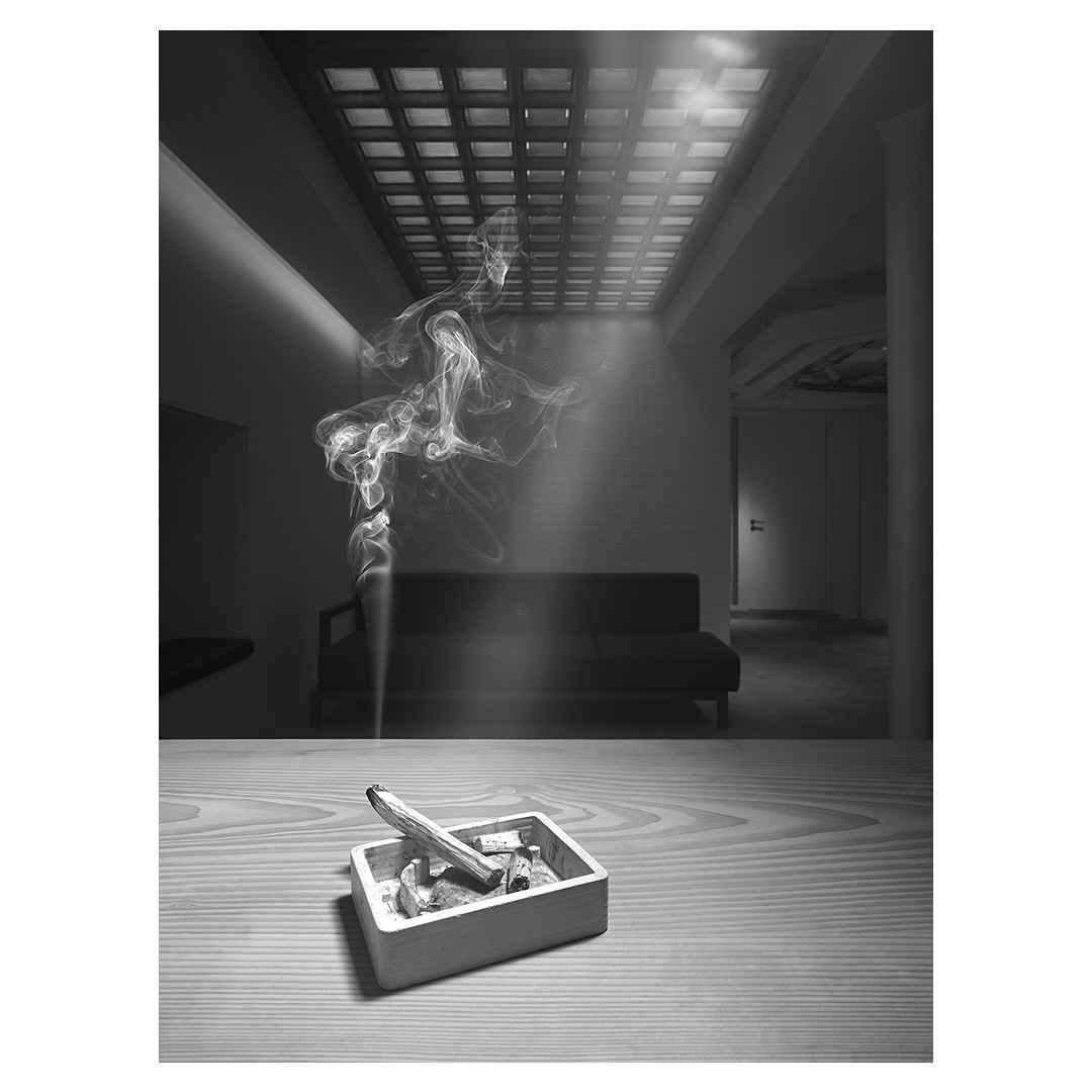 Indoor shot in monochrome of an ashtray with a smoking incense stick on a table, with wisps of smoke rising in the interior space.