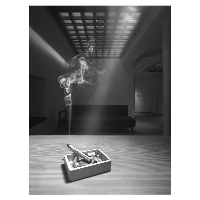 Indoor shot in monochrome of an ashtray with a smoking incense stick on a table, with wisps of smoke rising in the interior space.