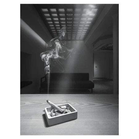 Indoor shot in monochrome of an ashtray with a smoking incense stick on a table, with wisps of smoke rising in the interior space.
