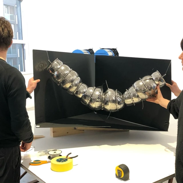 Two people hold a large black display with a silver, caterpillar-like sculpture made of metal colanders attached, studio lit.
