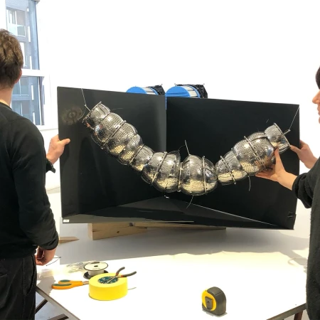 Two people hold a large black display with a silver, caterpillar-like sculpture made of metal colanders attached, studio lit.