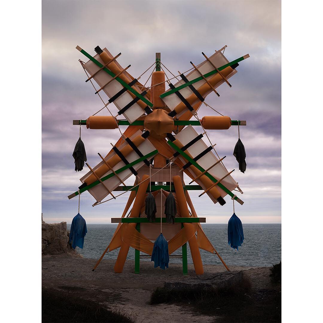 Artistic windmill sculpture on a rocky coastline under a cloudy sky, featuring textile hangings, orange and green framework, and ocean backdrop.