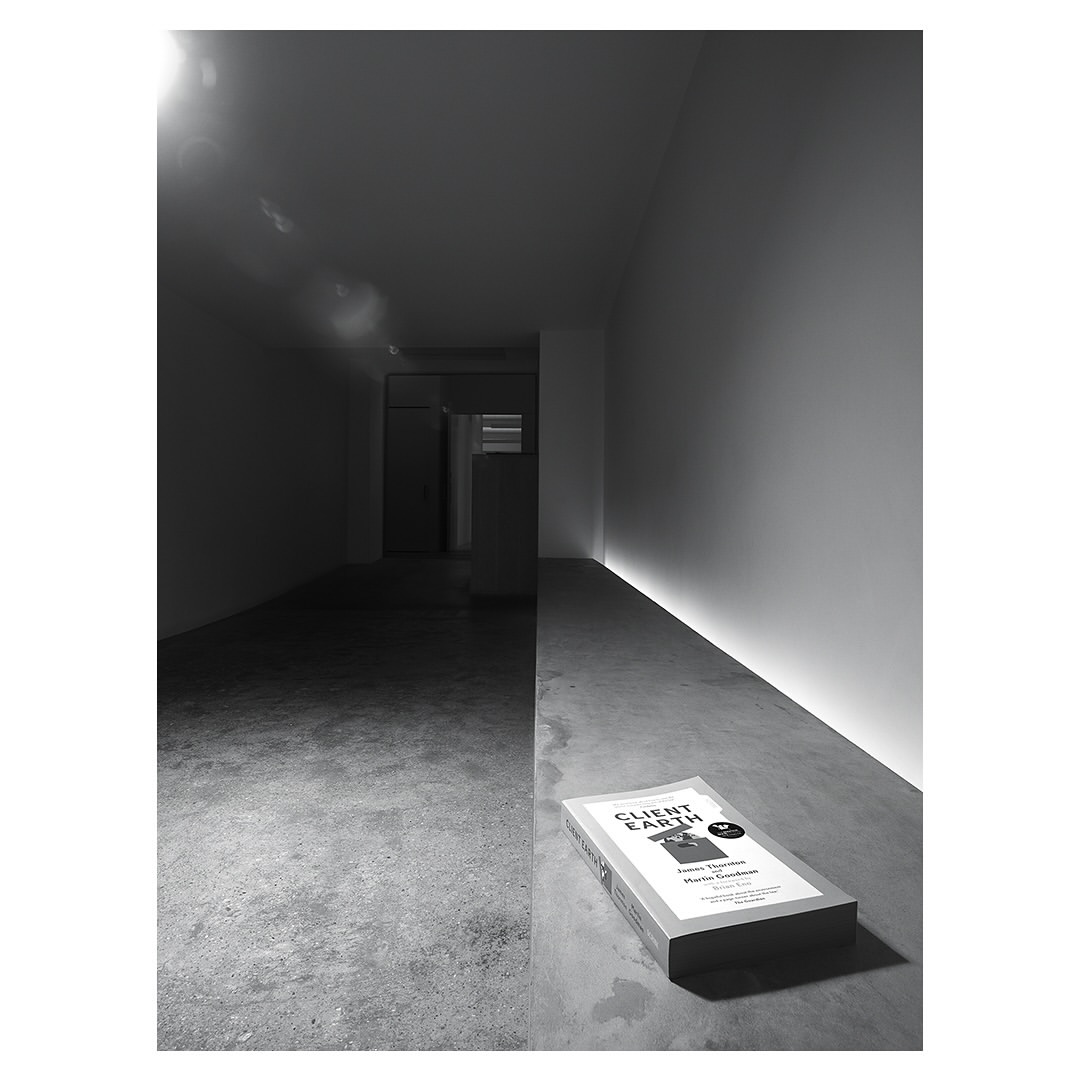 Monochromatic shot of a book titled 'Client Earth' laying on a concrete floor with a hallway in the background and subtle light.
