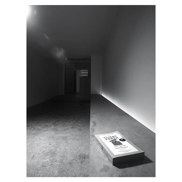 Monochromatic shot of a book titled 'Client Earth' laying on a concrete floor with a hallway in the background and subtle light.