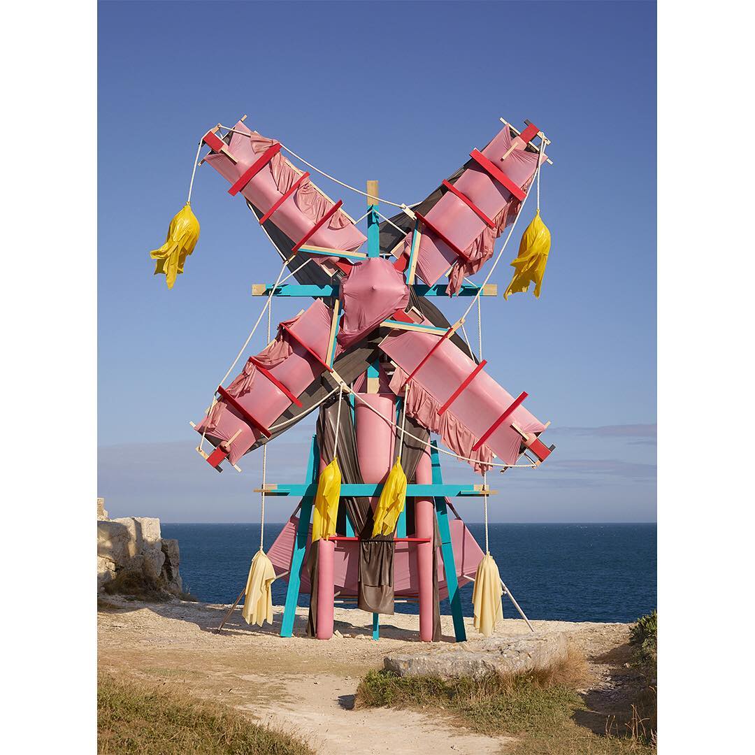 Outdoor view of a whimsical windmill with pink and turquoise wooden supports, adorned with pink fabric and yellow fringe.