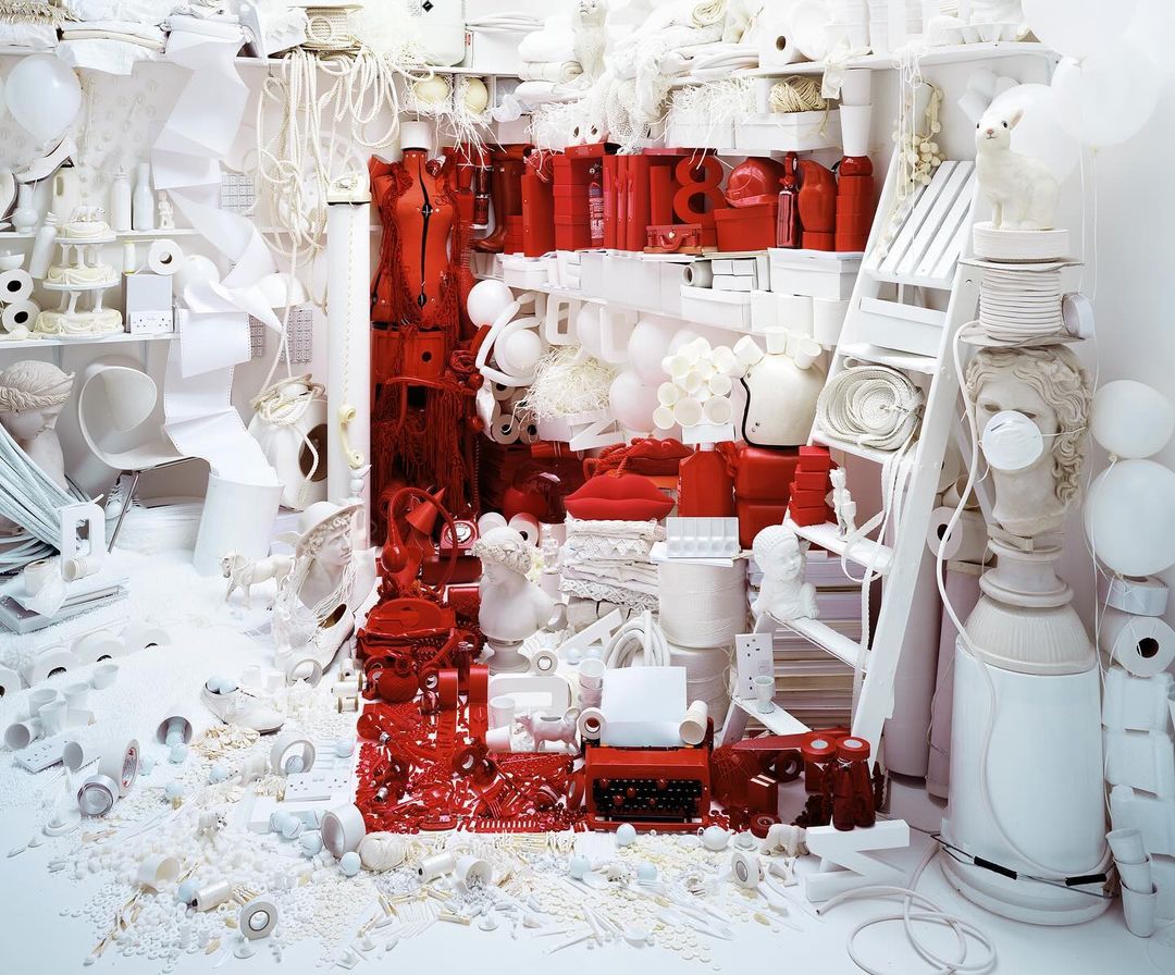 Surreal still life with white and red objects filling the frame, including a dress form, typewriter, busts, and scattered elements in a detailed scene.