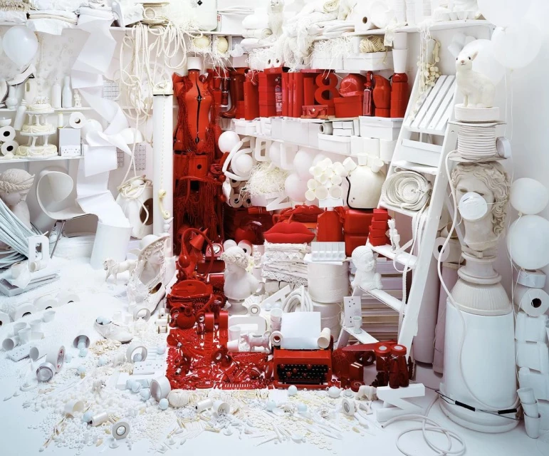 Surreal still life with white and red objects filling the frame, including a dress form, typewriter, busts, and scattered elements in a detailed scene.