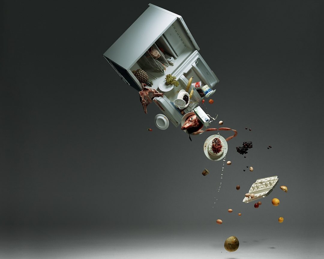Conceptual photography of a tipped-over refrigerator with an assortment of food items floating out on a gray background.