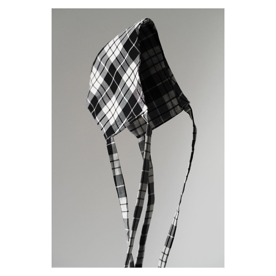 Black and white studio photograph of a plaid bonnet. The bonnet is displayed on a solid gray background, creating a minimalist fashion image.