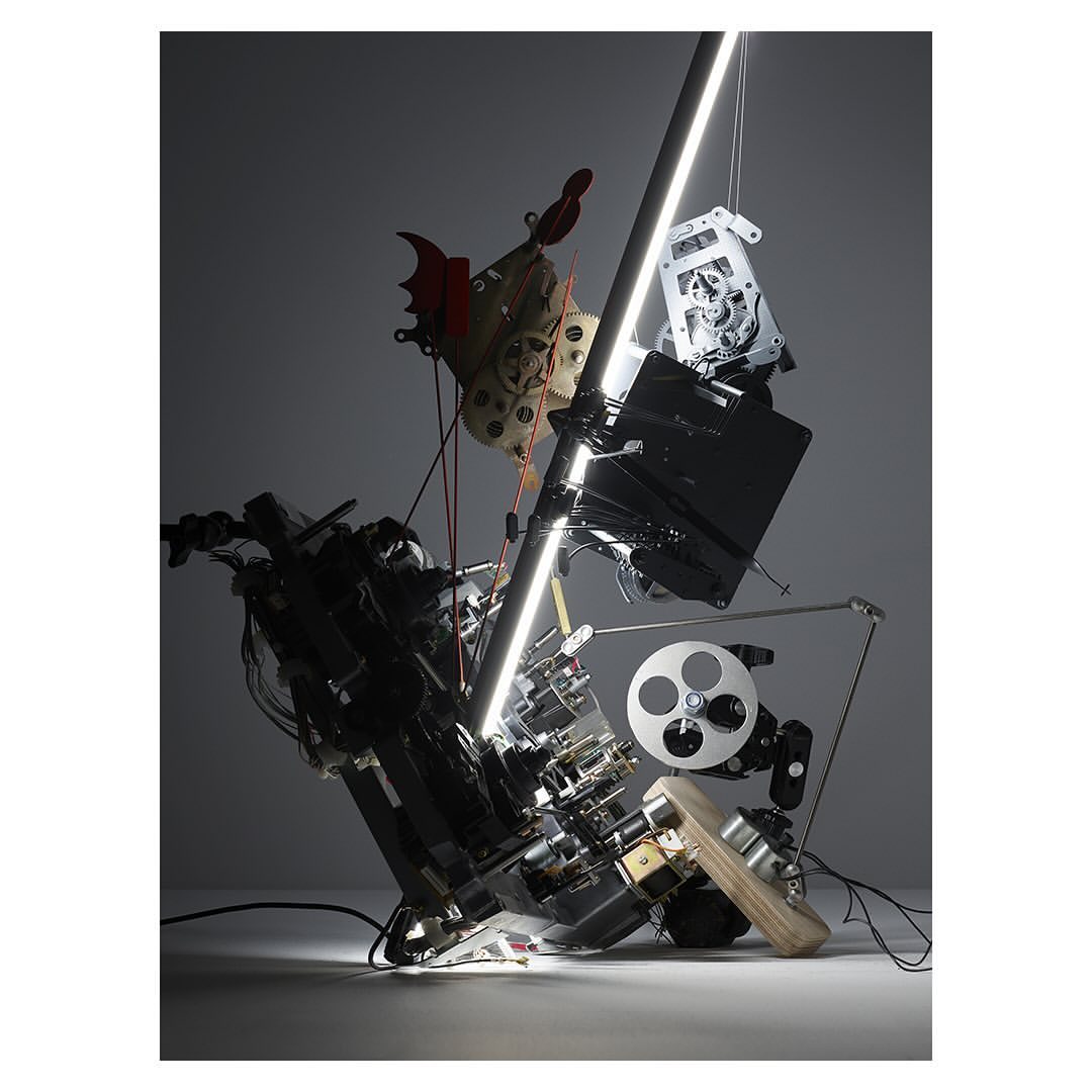 Abstract arrangement of electronic components, a chicken mask, and a film reel, illuminated by a vertical light, creating a complex, sculptural composition.