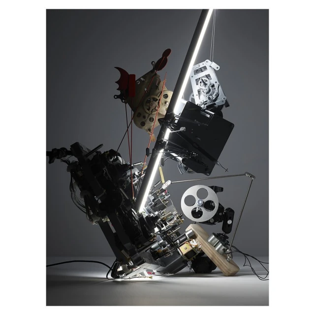 Abstract arrangement of electronic components, a chicken mask, and a film reel, illuminated by a vertical light, creating a complex, sculptural composition.