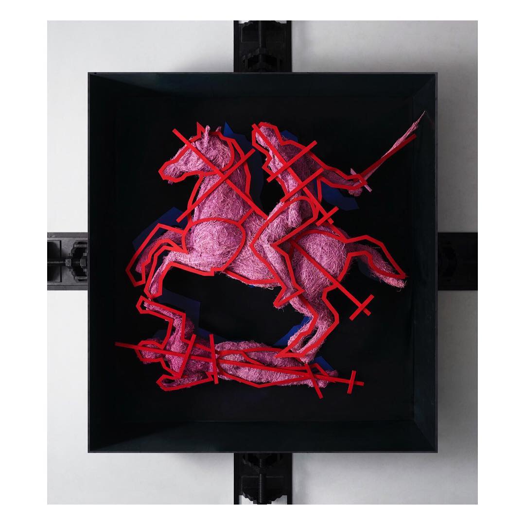 Art installation in a black frame featuring a pink thread sculpture of a horse and rider slaying a prone figure with a sword.