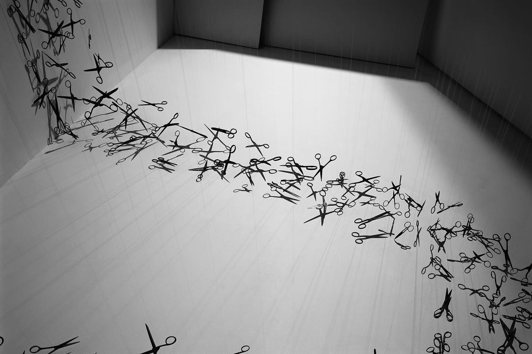 Monochromatic art installation featuring a cascade of scissors suspended from the ceiling in a white corner room, creating a surreal display.