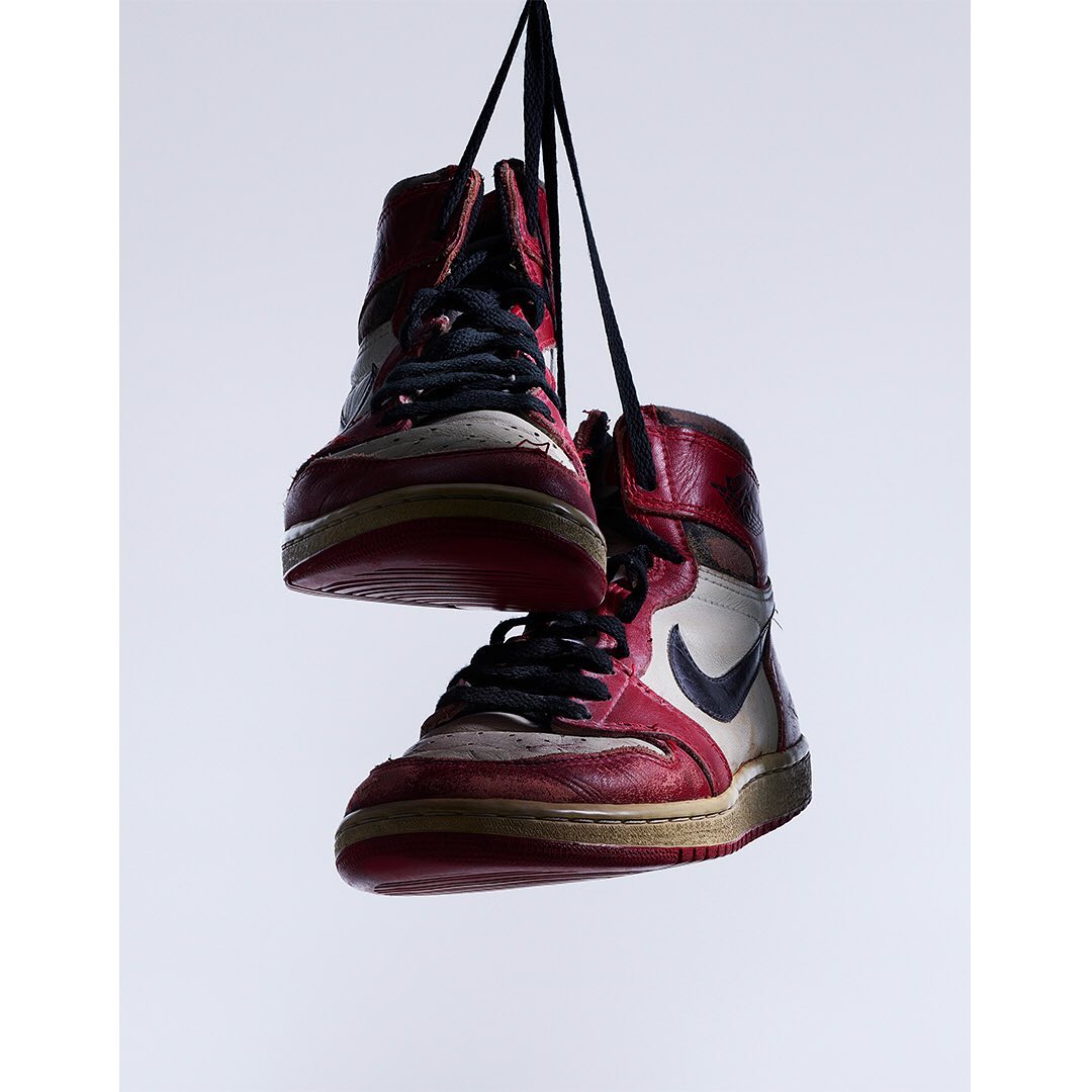 Two red and white high-top sneakers are suspended by their black laces against a gradient white backdrop in a product shot.