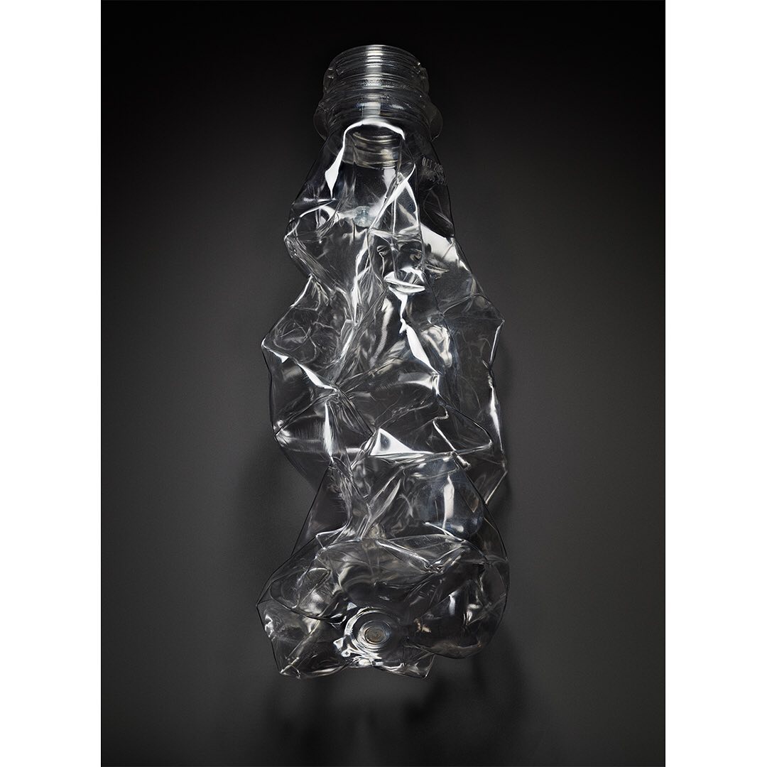 Still life studio shot of a crumpled clear plastic bottle, angled on a dark gray background with diffused lighting and soft shadows.
