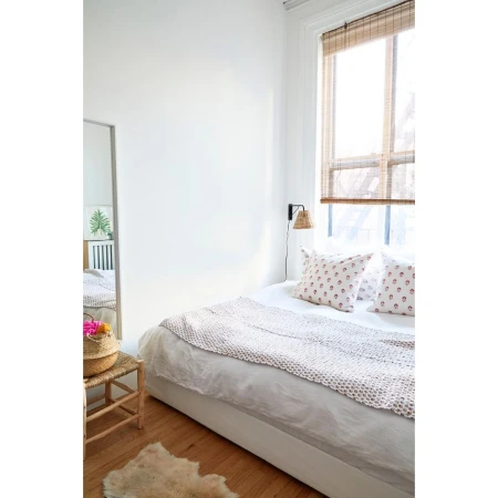 Bright, minimalist bedroom interior with a white bed, geometric throw, patterned pillows, rattan lamp, and natural window shades.