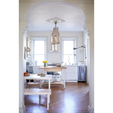 Bright, minimalist kitchen with white cabinets, hardwood floors, light wood table and bench, pendant light, and large windows.