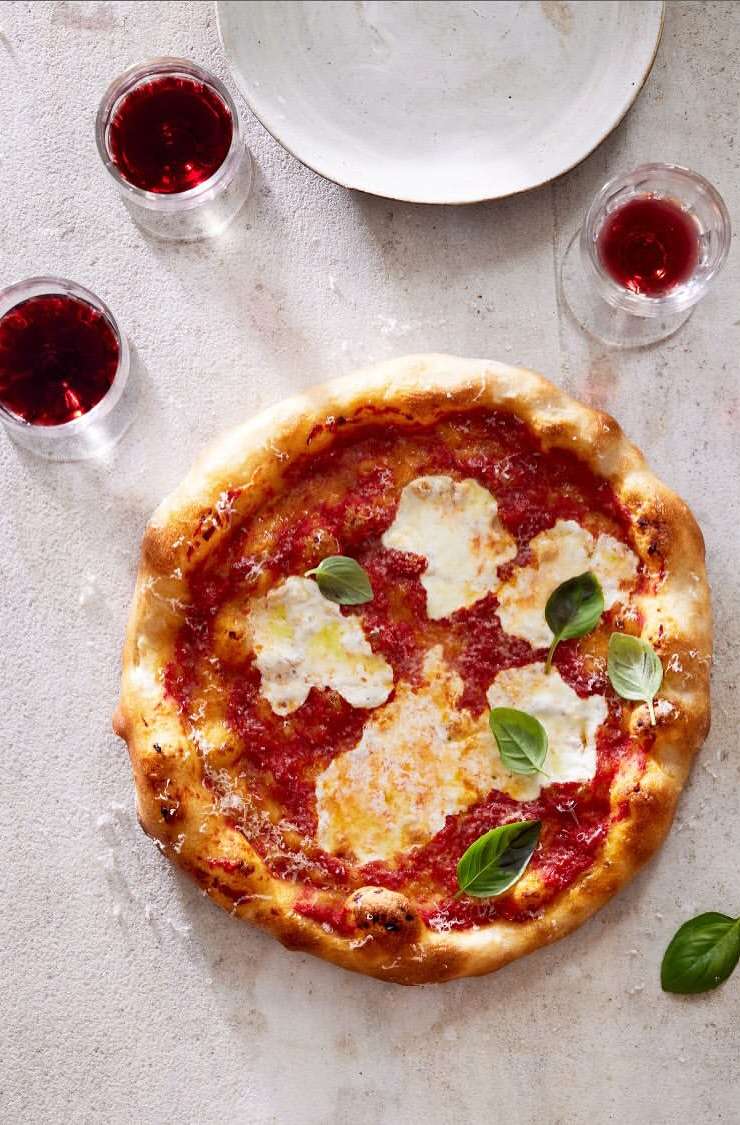 Overhead view of Margherita pizza with tomato sauce, mozzarella cheese, basil leaves, glasses of red wine, and a white plate.
