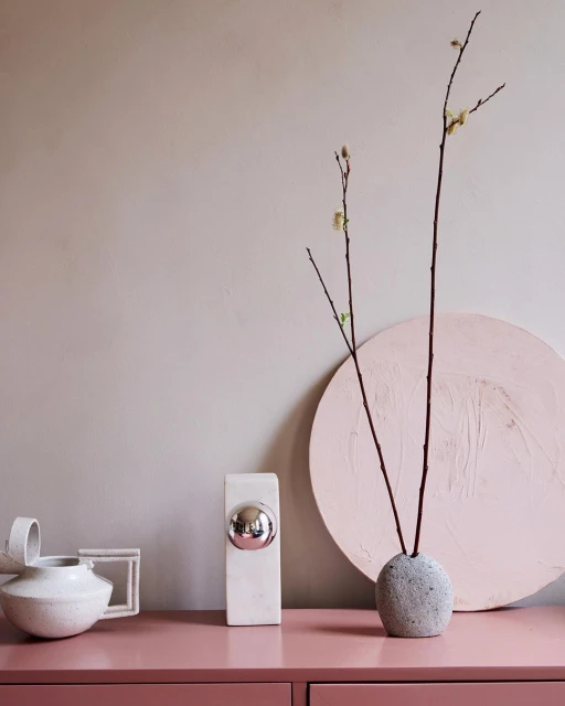 Still life of home decor including minimalist vases, dried branches, and a pink dresser against a neutral wall, indoor setting.