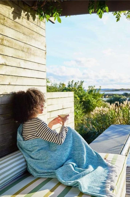 Woman wrapped in a blue blanket sits outdoors on a sunny day with a cup in hand, overlooking water and greenery, in a relaxed scene.