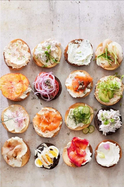Overhead shot of 16 bagel halves, each uniquely topped with cream cheese and various savory toppings on a neutral-toned background.