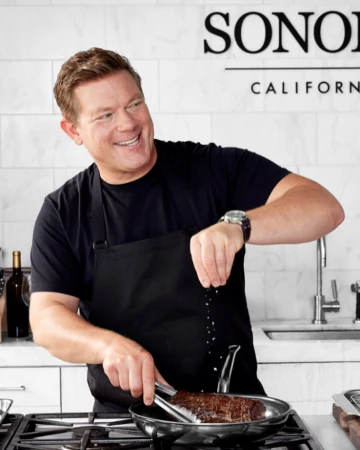 Chef salts a steak cooking in a pan on a stove in a kitchen, wearing a black shirt and apron in front of a white marble wall.