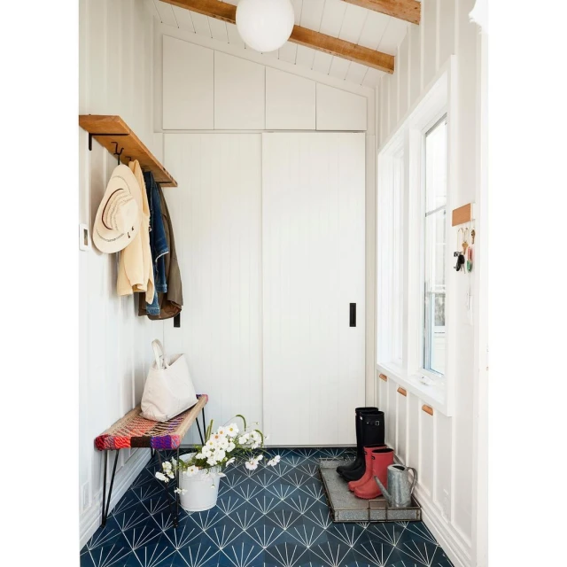 Bright, airy mudroom with patterned blue tile floor, white walls, storage, colorful bench, flowers, boots, and metal watering can.