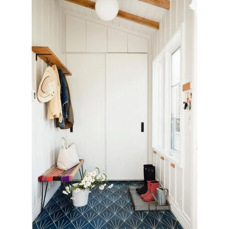 Bright, airy mudroom with patterned blue tile floor, white walls, storage, colorful bench, flowers, boots, and metal watering can.