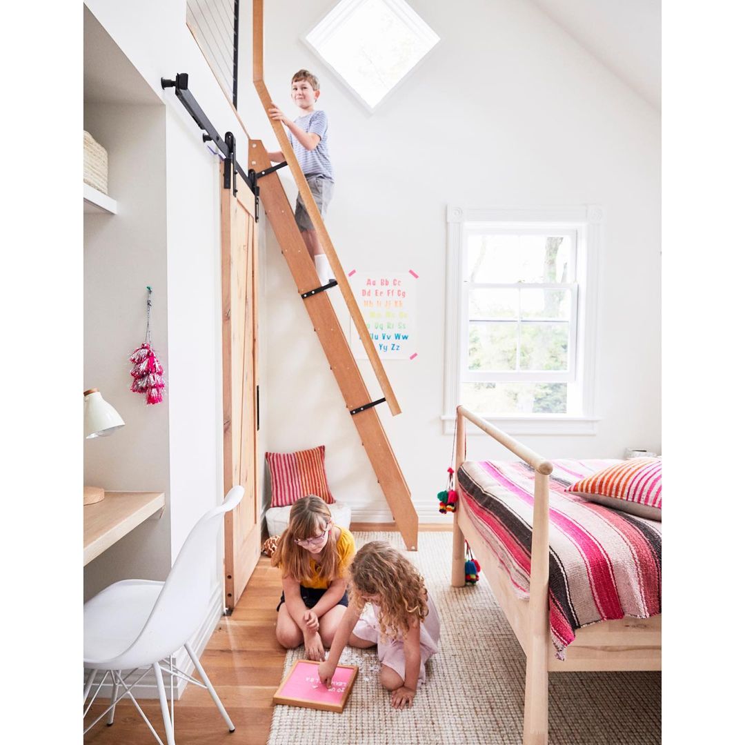 Three children playing in a brightly lit bedroom with a ladder leading to a loft, alongside a bed, desk, and alphabet chart.