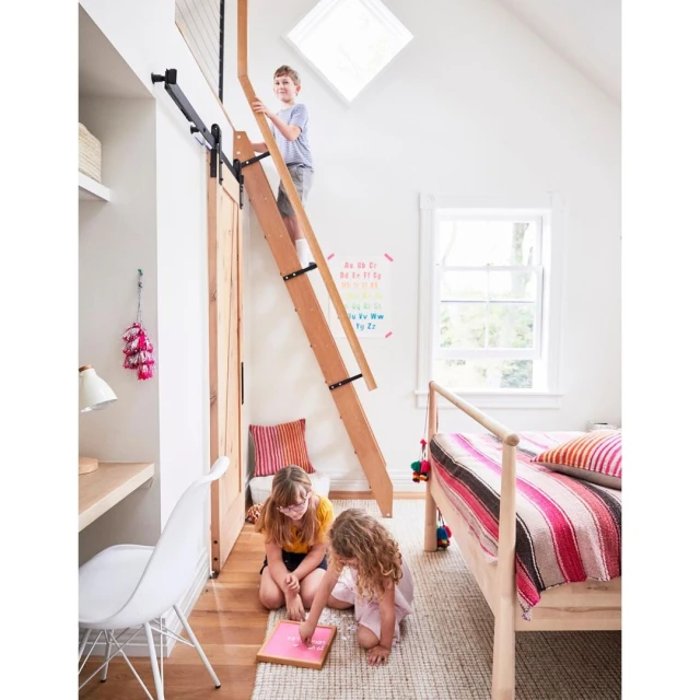 Three children playing in a brightly lit bedroom with a ladder leading to a loft, alongside a bed, desk, and alphabet chart.