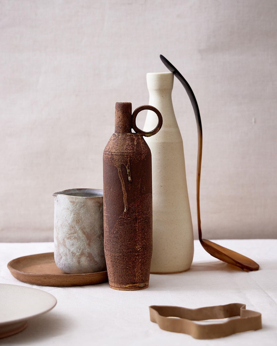 Still life with a rusty brown bottle vase, white ceramic vases, a wooden spoon, and a cookie cutter on a white linen tablecloth.