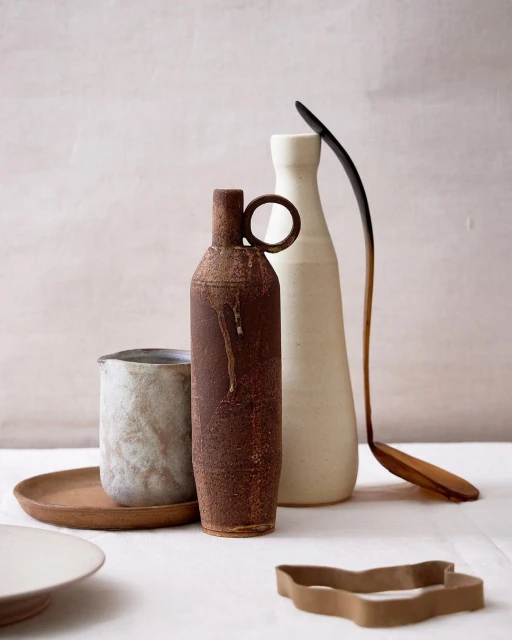 Still life with a rusty brown bottle vase, white ceramic vases, a wooden spoon, and a cookie cutter on a white linen tablecloth.