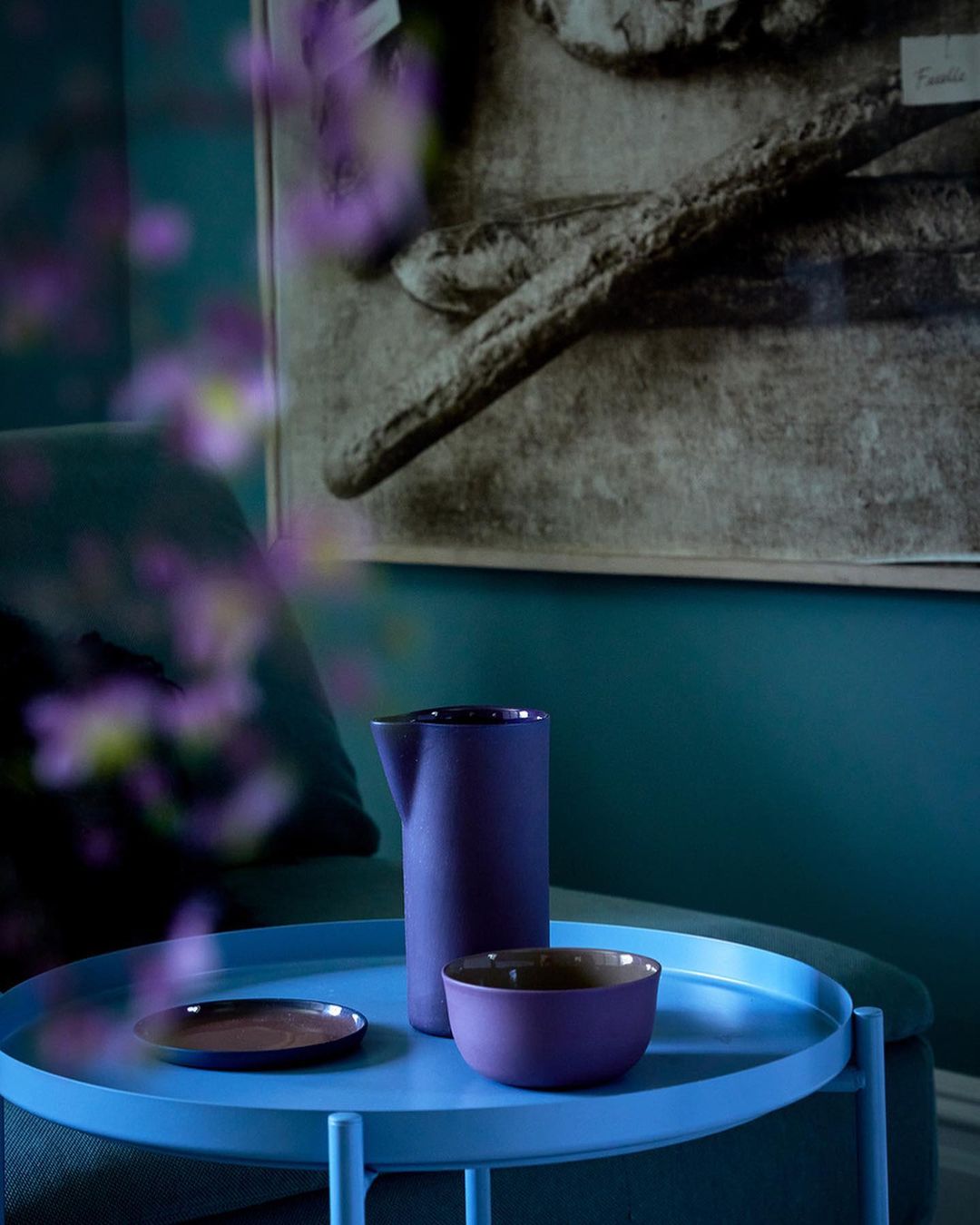 Still life of a purple pitcher, bowl, and plate on a blue table in front of teal wall with framed bread art.