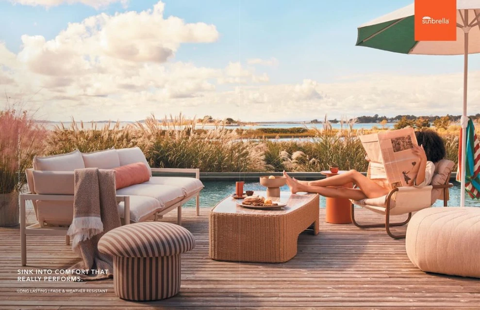 Outdoor patio scene featuring a woman reading, comfortable furniture arrangement, an umbrella, and a distant seascape view.