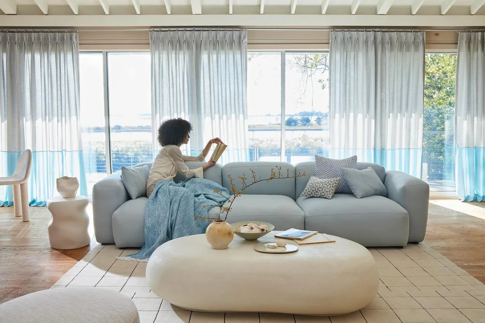 Woman reading a book on a blue sofa with pillows and a throw blanket in a living room with large windows and blue curtains.