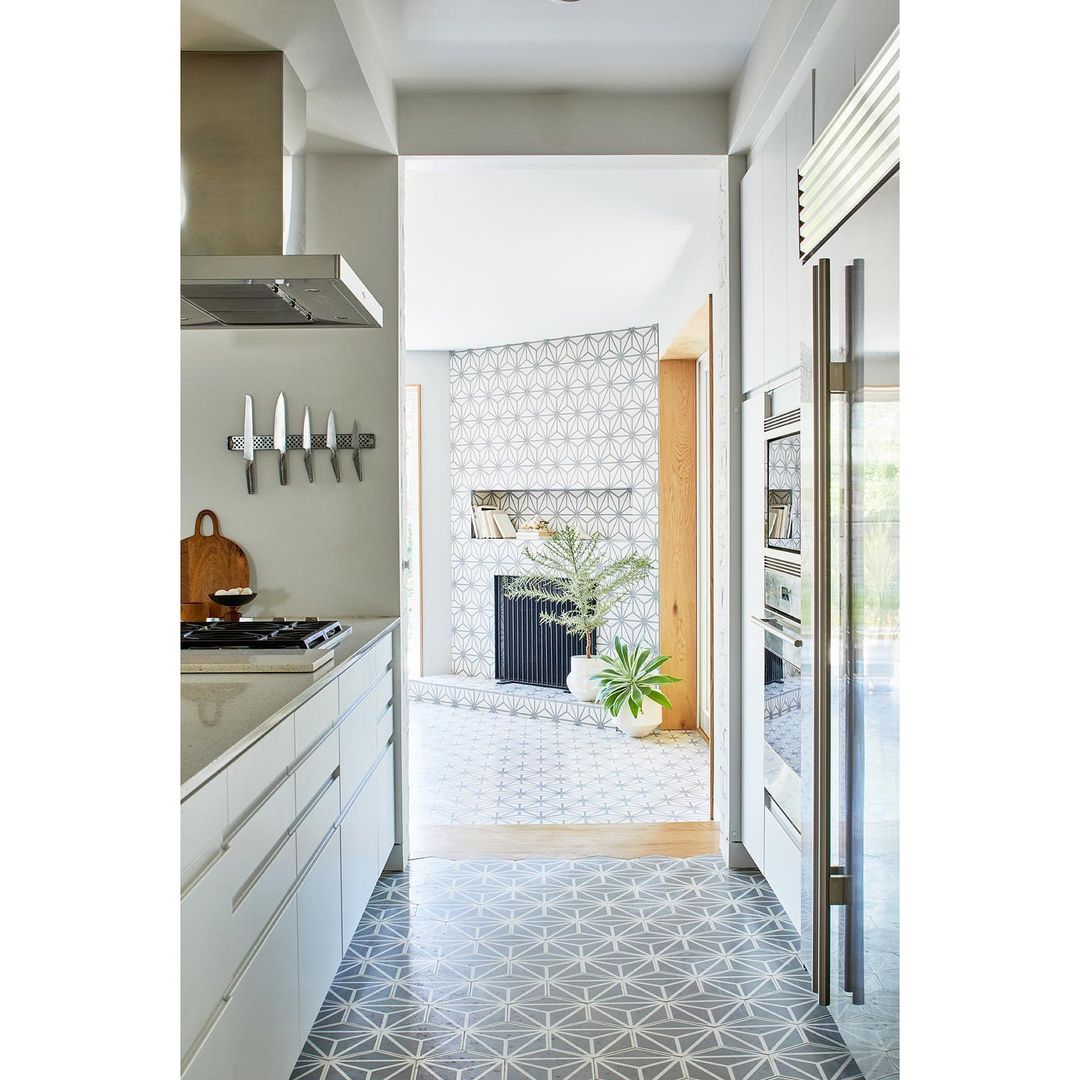 Modern kitchen interior with white cabinets, stainless steel appliances, geometric patterned floor tiles, and a view into a living area.