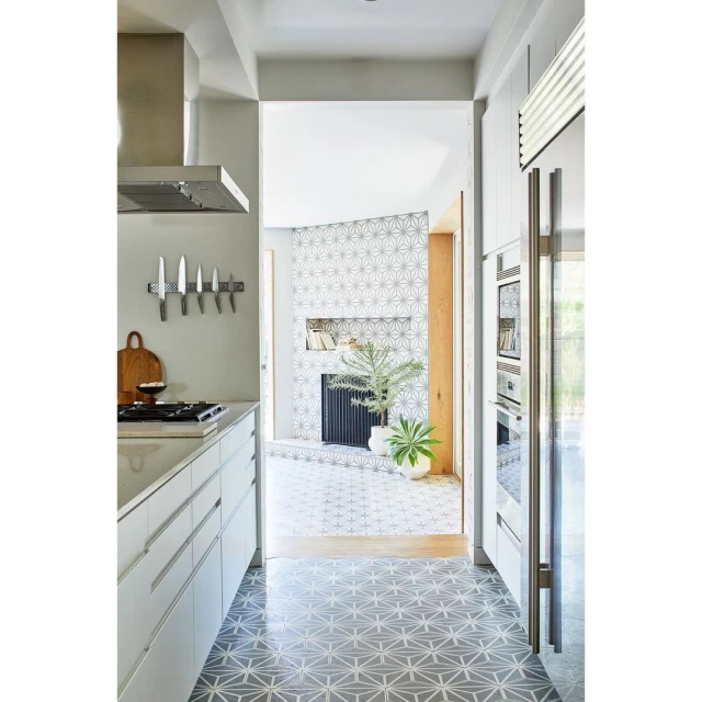 Modern kitchen interior with white cabinets, stainless steel appliances, geometric patterned floor tiles, and a view into a living area.