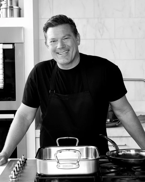 Monochrome portrait of a smiling adult male in a black apron standing in a kitchen with stainless steel cookware on the stovetop.