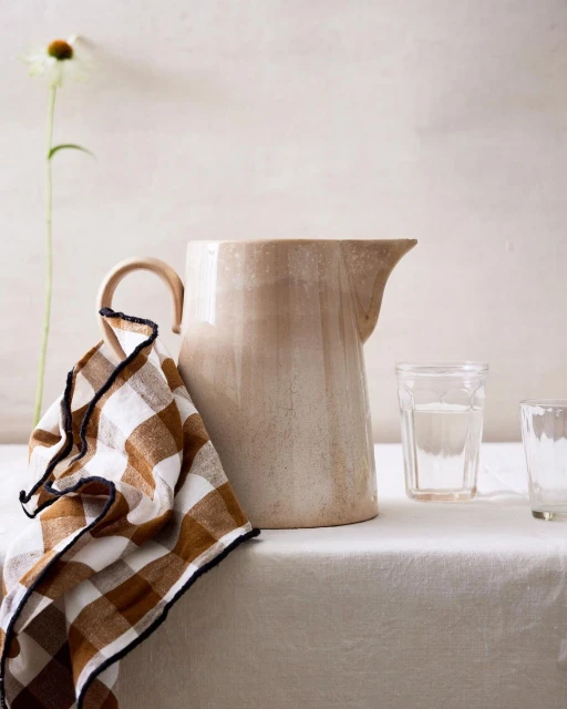 Still life photograph featuring a beige ceramic pitcher, two clear glasses, a checkered brown and white towel, and a white flower.